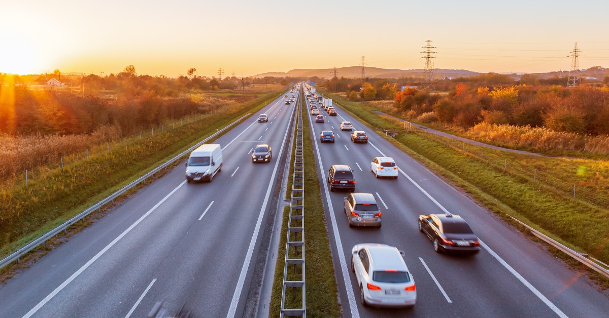 Two sides of a highway with cars traveling in opposite directions. Grass and trees line each side of the highway.