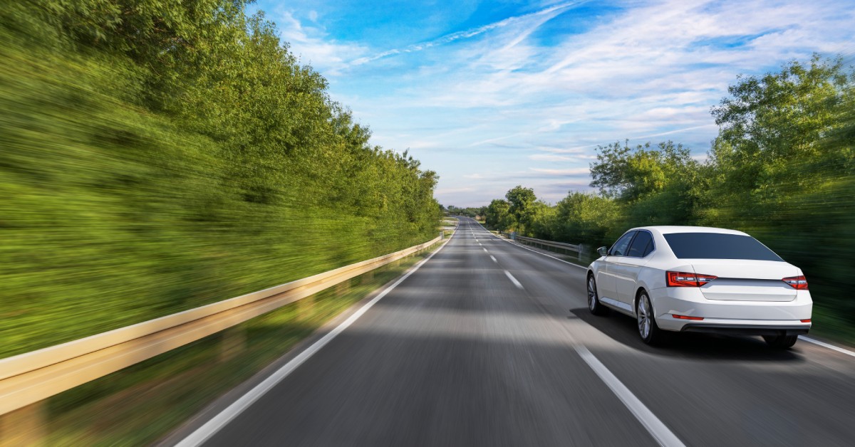 The back of a white sedan driving down a rural road with trees blurring past it. The sky is bright blue above.