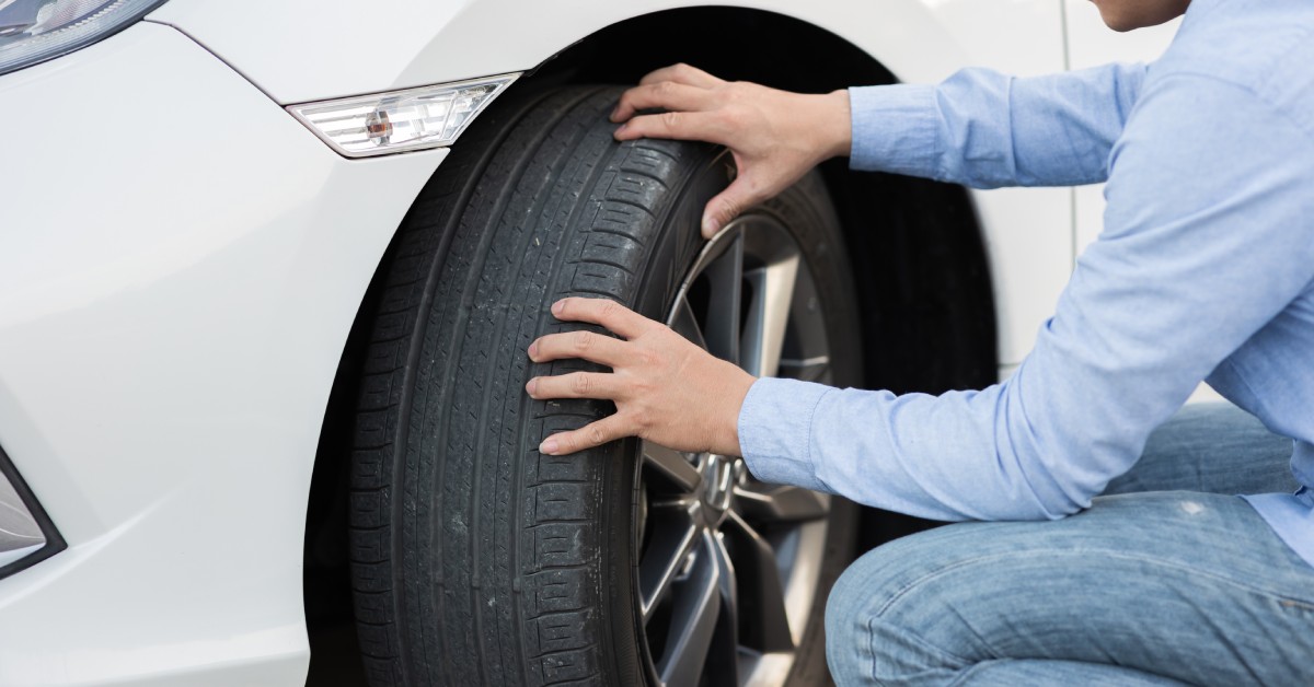 A person kneeling next to a car with both of their hands on the side of the tire. The tire is on the front of the white car.
