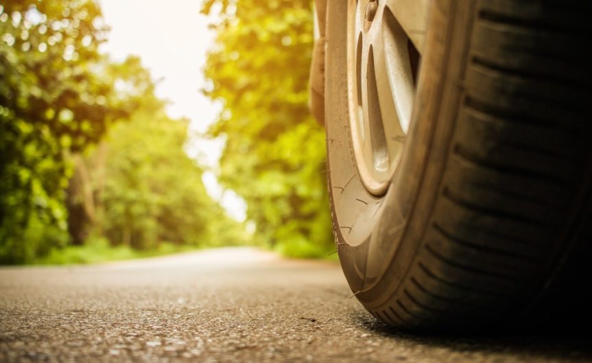 The wheel of a parked car on an asphalt road. Behind the tire is a clear road surrounded by green trees.