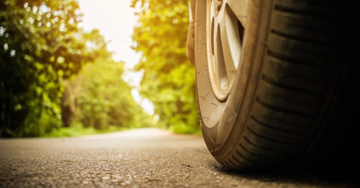 The wheel of a parked car on an asphalt road. Behind the tire is a clear road surrounded by green trees.