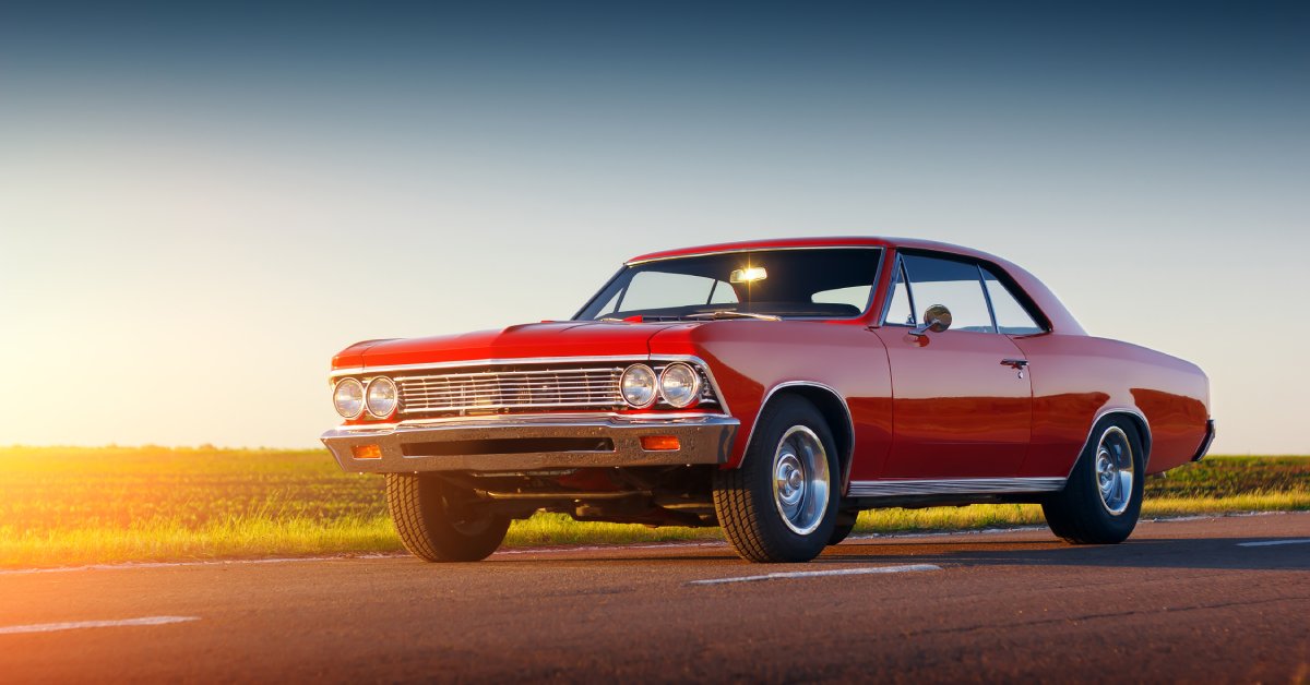 A retro red car sitting on the open road with the sun setting behind it. There is also a green field behind the road.