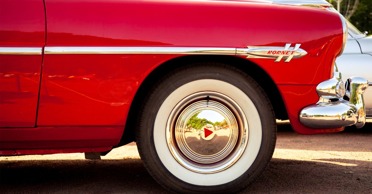 The front side of a red car with a vintage design parked on asphalt. The car's tire has a white outline.