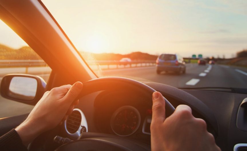 A driver's view of a windshield with the driver's hands on the steering wheel. They are driving on a highway during sunset.