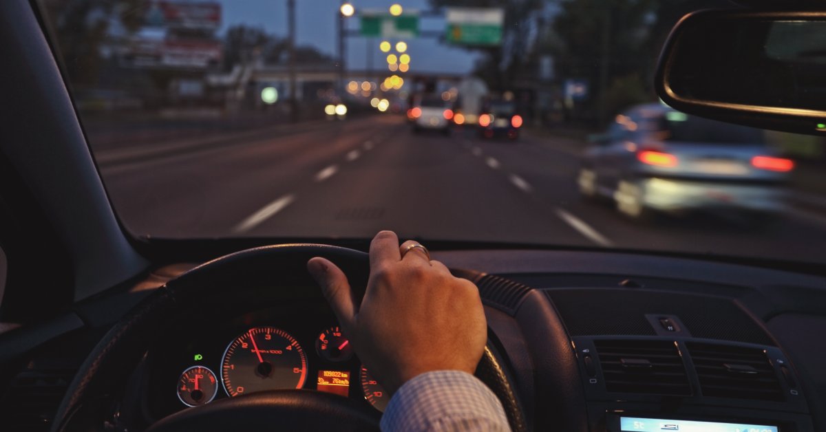 A person has one hand on a vehicle's steering wheel as they drive down a busy highway. The view is from the driver's perspective.