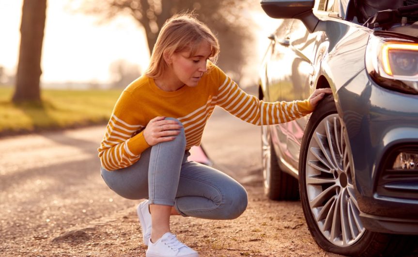 A woman on the side of the road with her vehicle. She is kneeling and looking at her tire for any problems.