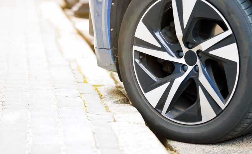 A car tire is partially on the curb of a sidewalk. The bottom bumper of the car and the entire wheel are visible.