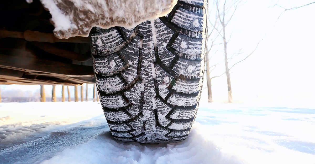 A rear view of a vehicle's winter tire with spikes. The vehicle is on a snowy road, and the tire tread has snow in it.