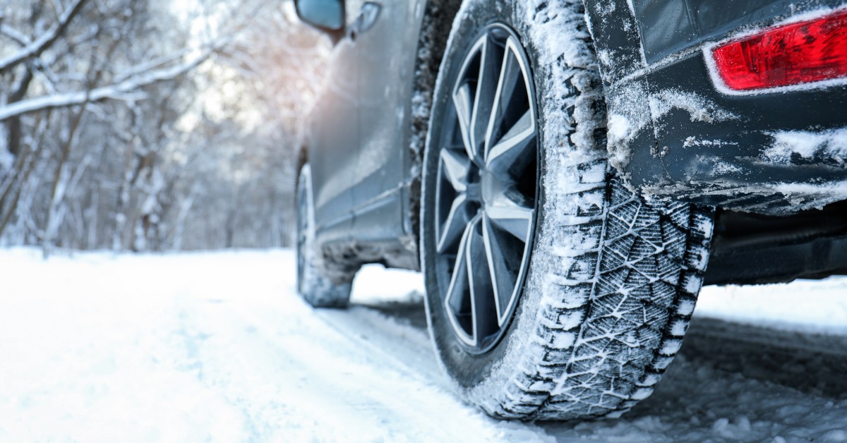 A rear view of a vehicle's winter tire with spikes. The vehicle is on a snowy road, and the tire tread has snow in it.