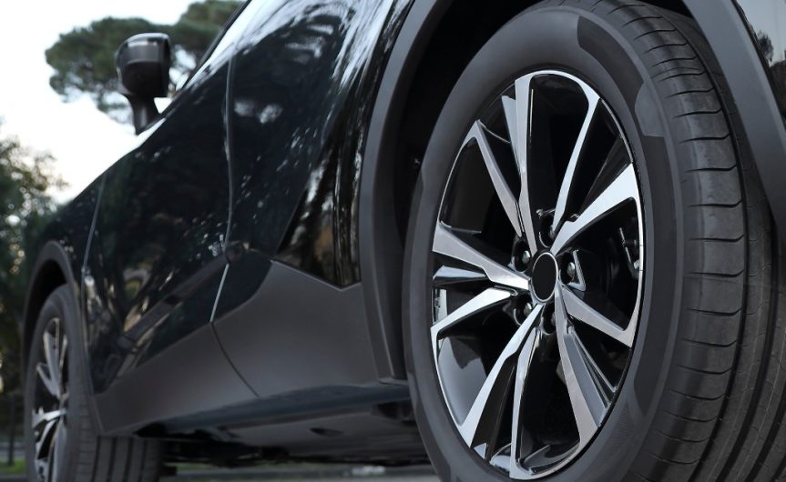 The side of a black car with clean front and back tires. The side of the vehicle reflects the trees around it.