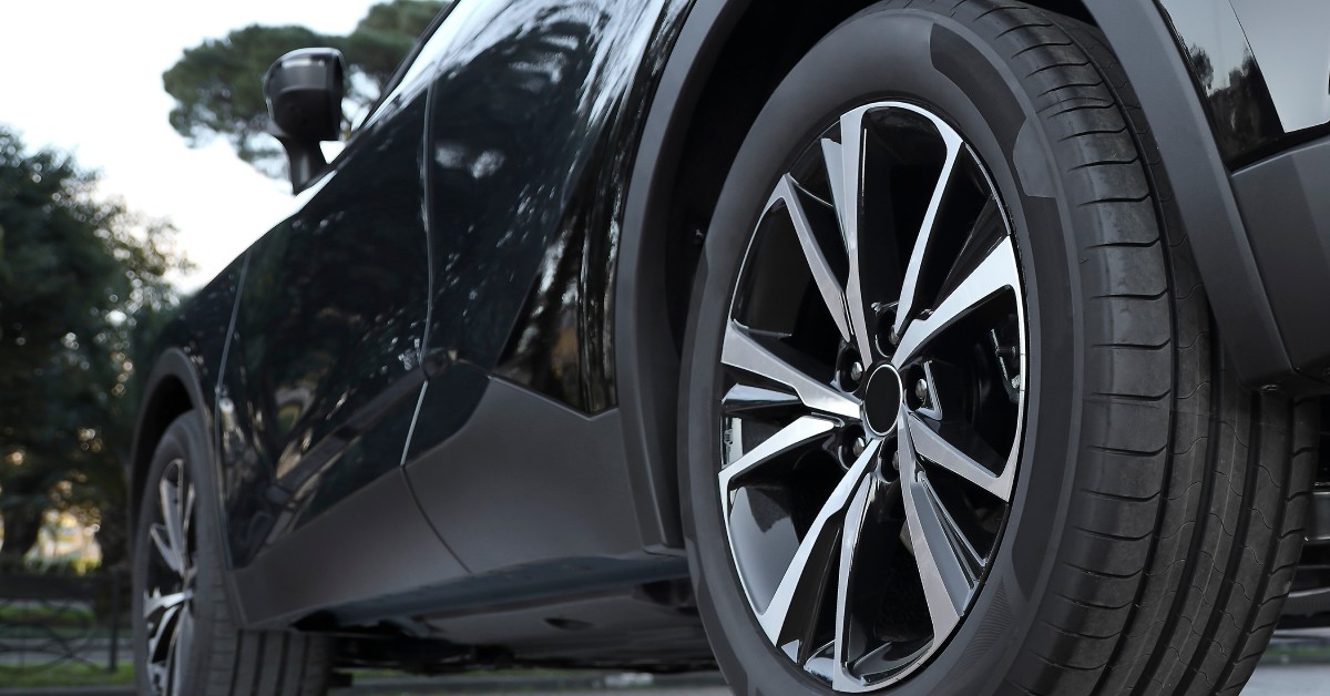 The side of a black car with clean front and back tires. The side of the vehicle reflects the trees around it.
