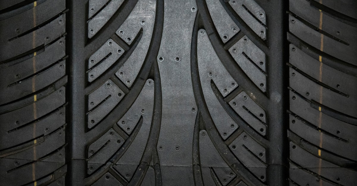 A close-up of a dark tire's tread containing intricate V-shaped grooves, circular indentations, and yellow vertical lines.