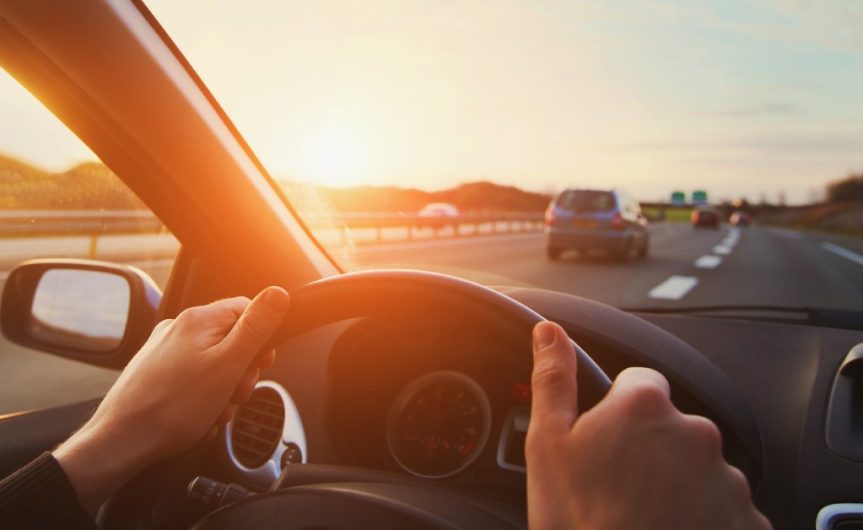 Behind the wheel of a car, a person's hands hold the wheel while on the highway. The sun is setting ahead of them.