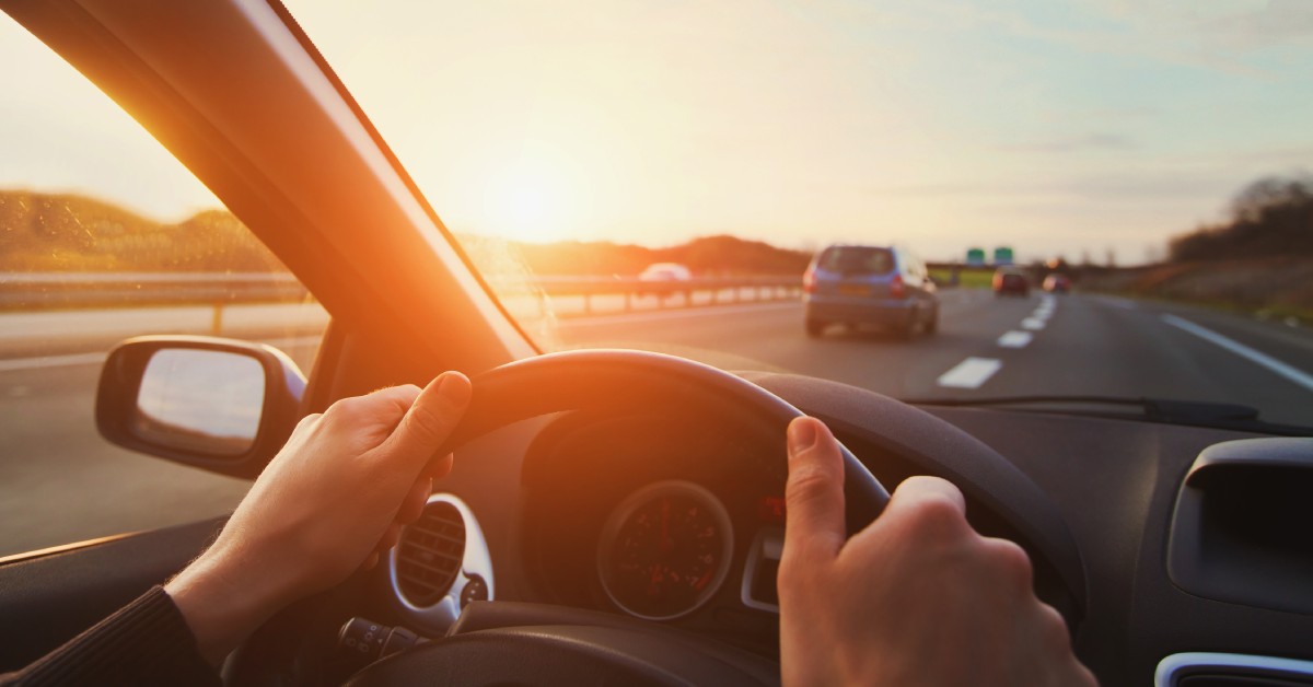 Behind the wheel of a car, a person's hands hold the wheel while on the highway. The sun is setting ahead of them.