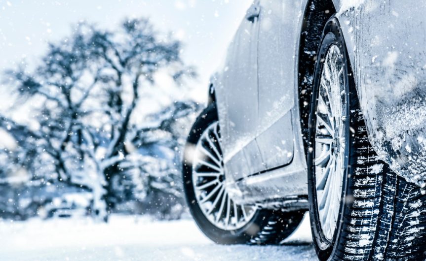 A car parked on a snowy road with trees in front of it. The vehicle's tires have snow stuck in the tread.