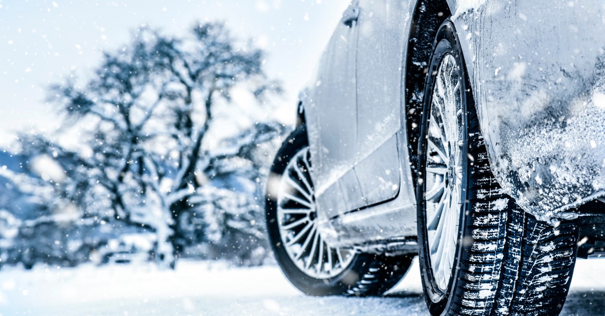 A car parked on a snowy road with trees in front of it. The vehicle's tires have snow stuck in the tread.