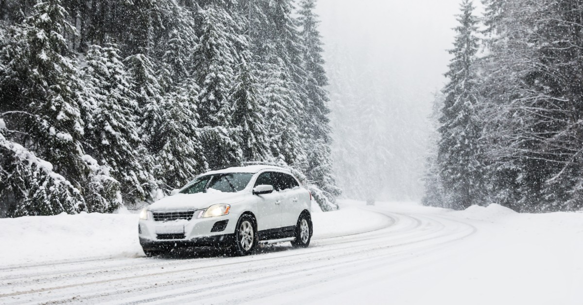 A white car driving on a snow-covered road with tall trees surrounding it. The vehicle also has snow on it.
