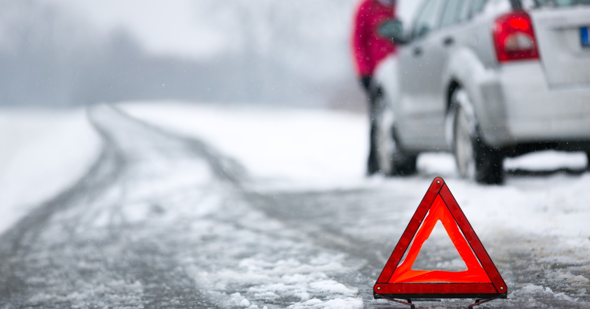 An emergency triangle sign on a snowy road with a car pulled over. A person is looking under the hood of the vehicle.