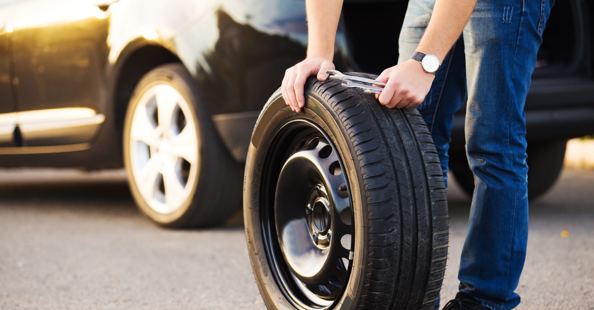 A man with his hands on a tire standing away from his parked car. He also has two wrenches in his hand.