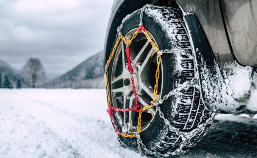 A tire has snow chains installed on it to help with driving on the snow. A snowy road lies ahead of the vehicle.