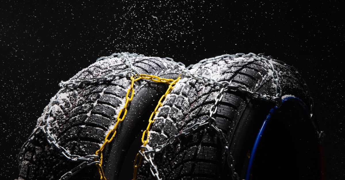 Two winter tires covered in snow are in front of a black background. Both tires have snow chains across the tread.