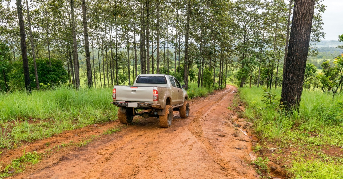 A silver truck drives down a wooded, muddy trail. The truck's tires are larger than average and caked in mud.