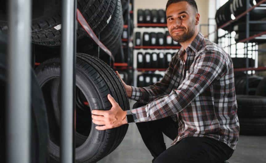 A young man picking up a tire from the bottom shelf of a rack. Behind him are other shelves of tires.