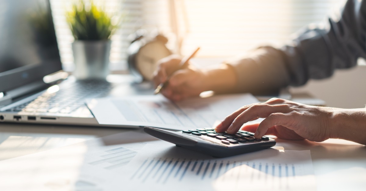 A person sitting at a table with a calculator and paper. There is also a laptop on the table under the paper.