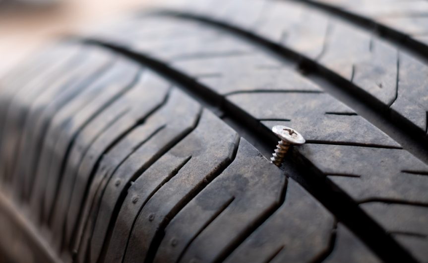 A car tire with a small puncture caused by a metal bolt. The bolt is wedged between the treads of the tire.