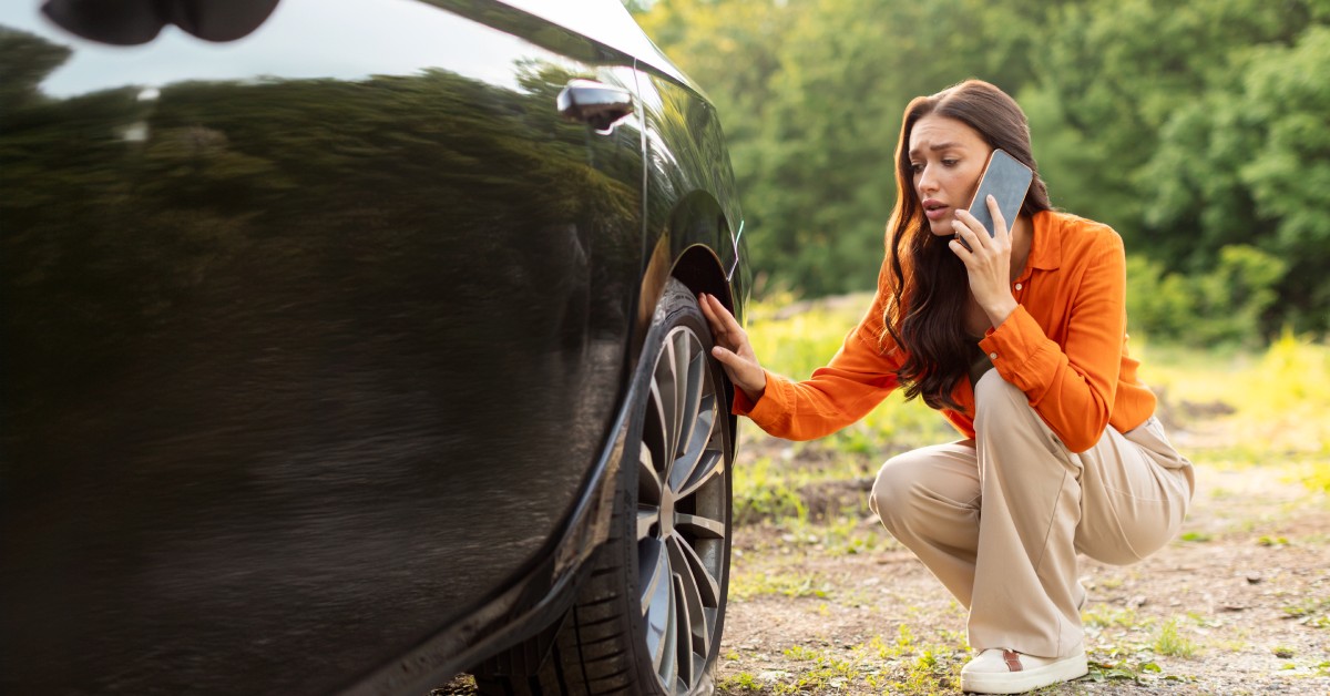 On a dirt road, a worried woman kneels beside her car, staring at the flat tire. She is on the phone with someone.