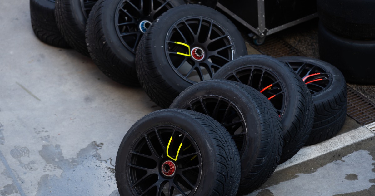 Eight black racing tires with yellow and red colored‑marked rims haphazardly rest on a concrete shop floor.