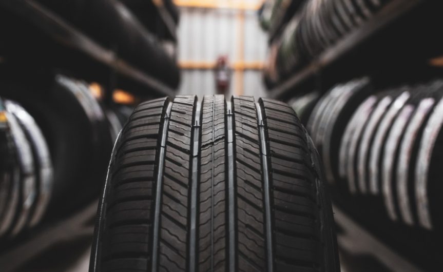 A close-up of the treads on a tire. Shelves of other new tires are blurred and on either side of the main tire.