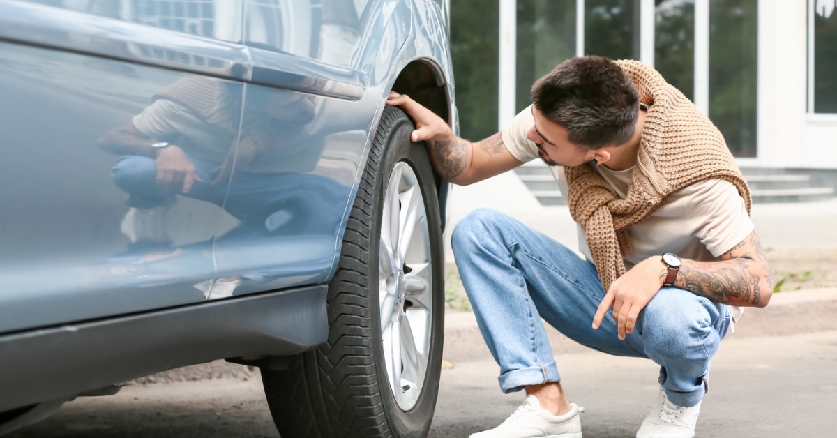 A young man in jeans, a white shirt, and white sneakers crouches down to examine the rear wheel tire of a blue car.