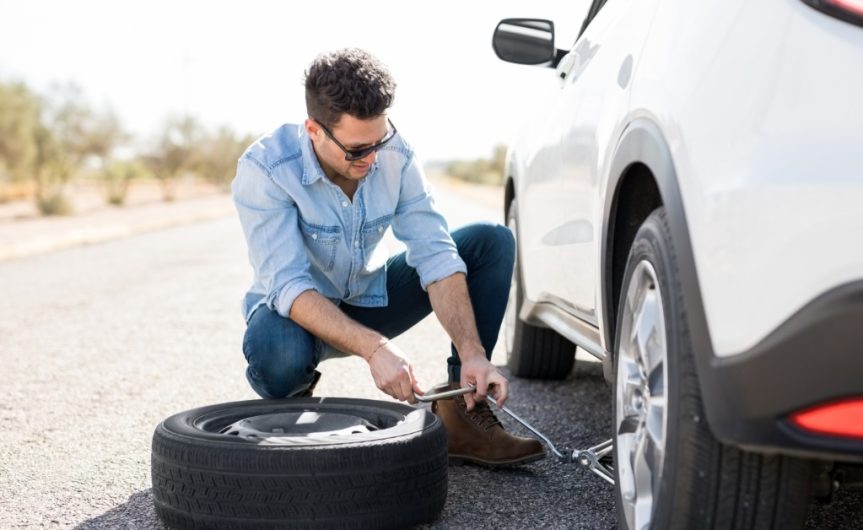 A man and a boy change a tire on a vehicle. The boy rolls a spare tire toward the man, loosening the lug nuts on the flat.