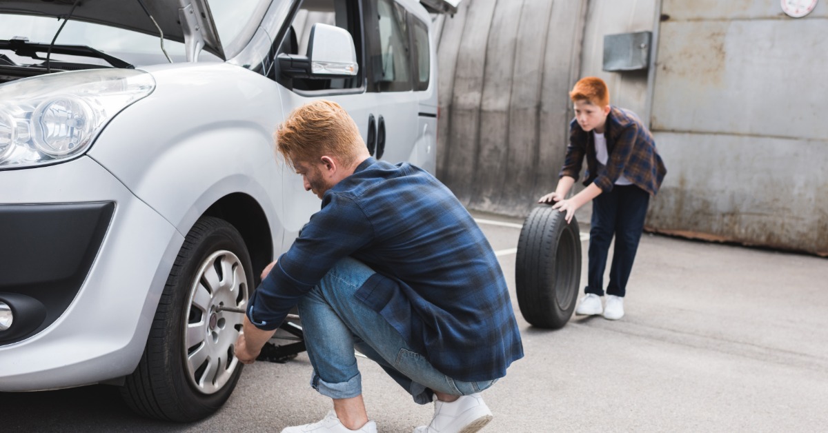 A man kneels next to the back tire of his car parked on the side of a road. He lifts the car on a jack with a spare tire next to him.