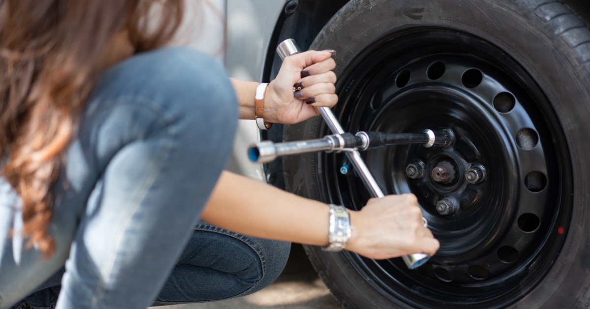 Someone kneels next to a spare tire fitted onto a vehicle. They use a lug wrench to tighten the nuts.