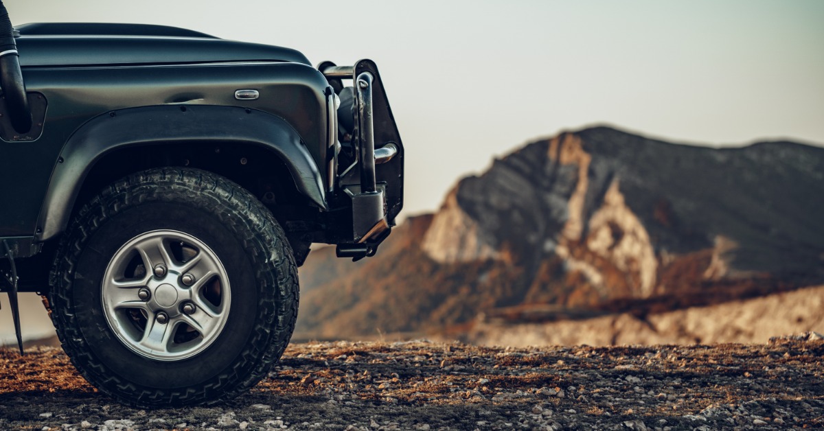 A close-up of the front end of a dark green Jeep with a grille guard, with mountains in the background.