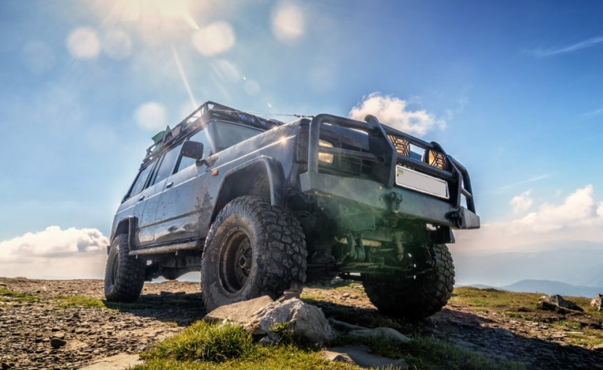 A ground-level view of a Jeep with a grille guard and off-road tires on top of a mountain on a clear, sunny day.