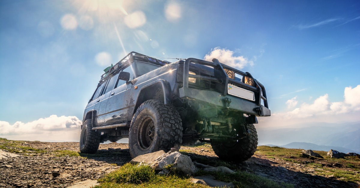 A ground-level view of a Jeep with a grille guard and off-road tires on top of a mountain on a clear, sunny day.