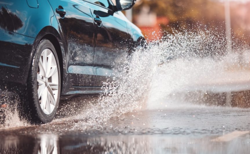 A black car drives through a large puddle during daylight. A spray of water shoots out from the vehicle's tires.