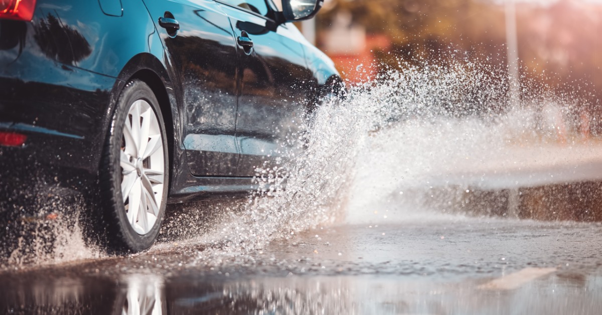 A black car drives through a large puddle during daylight. A spray of water shoots out from the vehicle's tires.