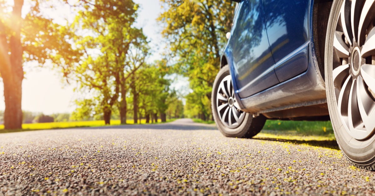 A blue car parked on the shoulder of a road with a close-up of the driver's side tires. The sun shines through some trees.