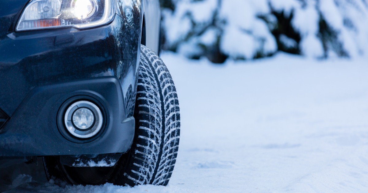 A close-up of the front of a black car with the wheel turned out. Snow covers the ground and the tire treads.