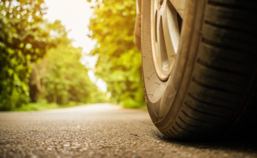 A car tire on a clear asphalt road with luscious green trees surrounding it. The sun is shining on the road.