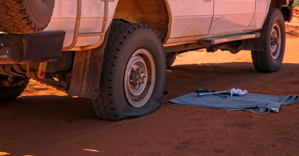 A car with a flat rear tire sitting on a sandy road. The sunlight is causing a shadow of the car over the sand.