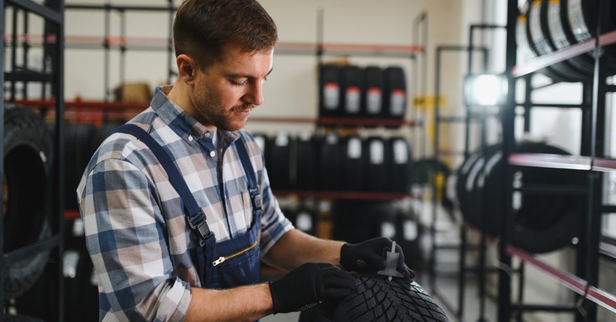 A man in a flannel shirt and overalls examining a new tire in a shop with a tire tread depth indicator.