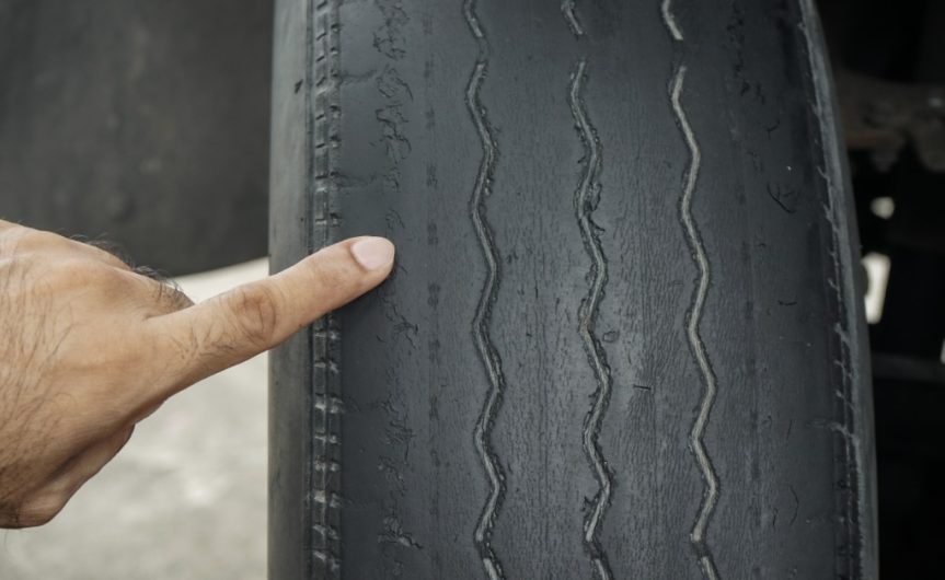 A close-up of a man's hand pointing with his index finger at the edge of a bald tire with the tread worn off.