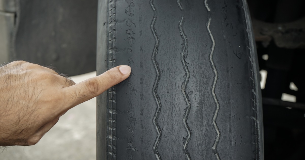 A close-up of a man's hand pointing with his index finger at the edge of a bald tire with the tread worn off.