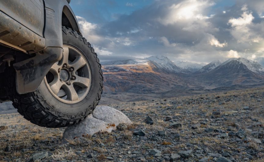 A close-up of the front tires of an off-road vehicle. The tire is posed on a rock with mountains in the background.