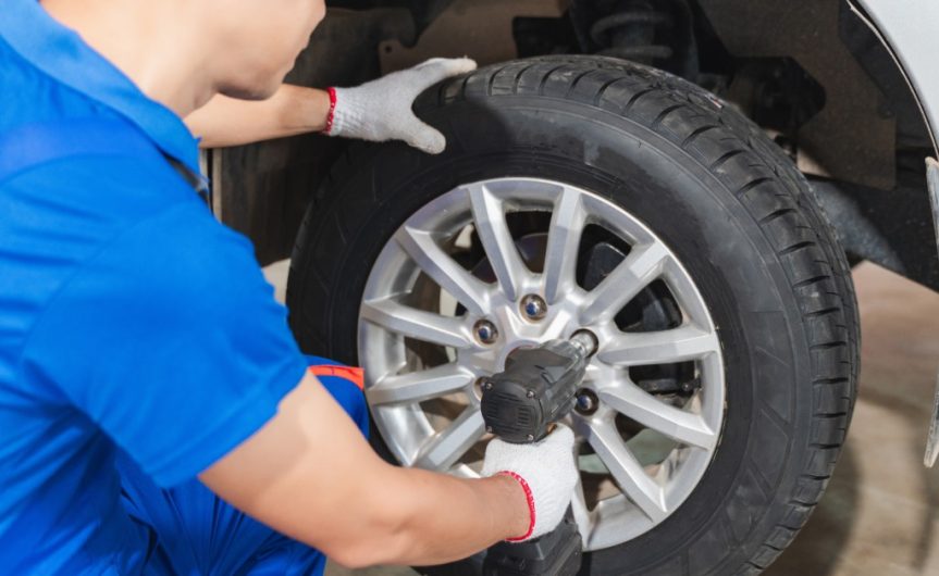 Looking over the shoulder of a mechanic as he uses a drill to tighten the lug nuts of a new wheel on a lifted car.